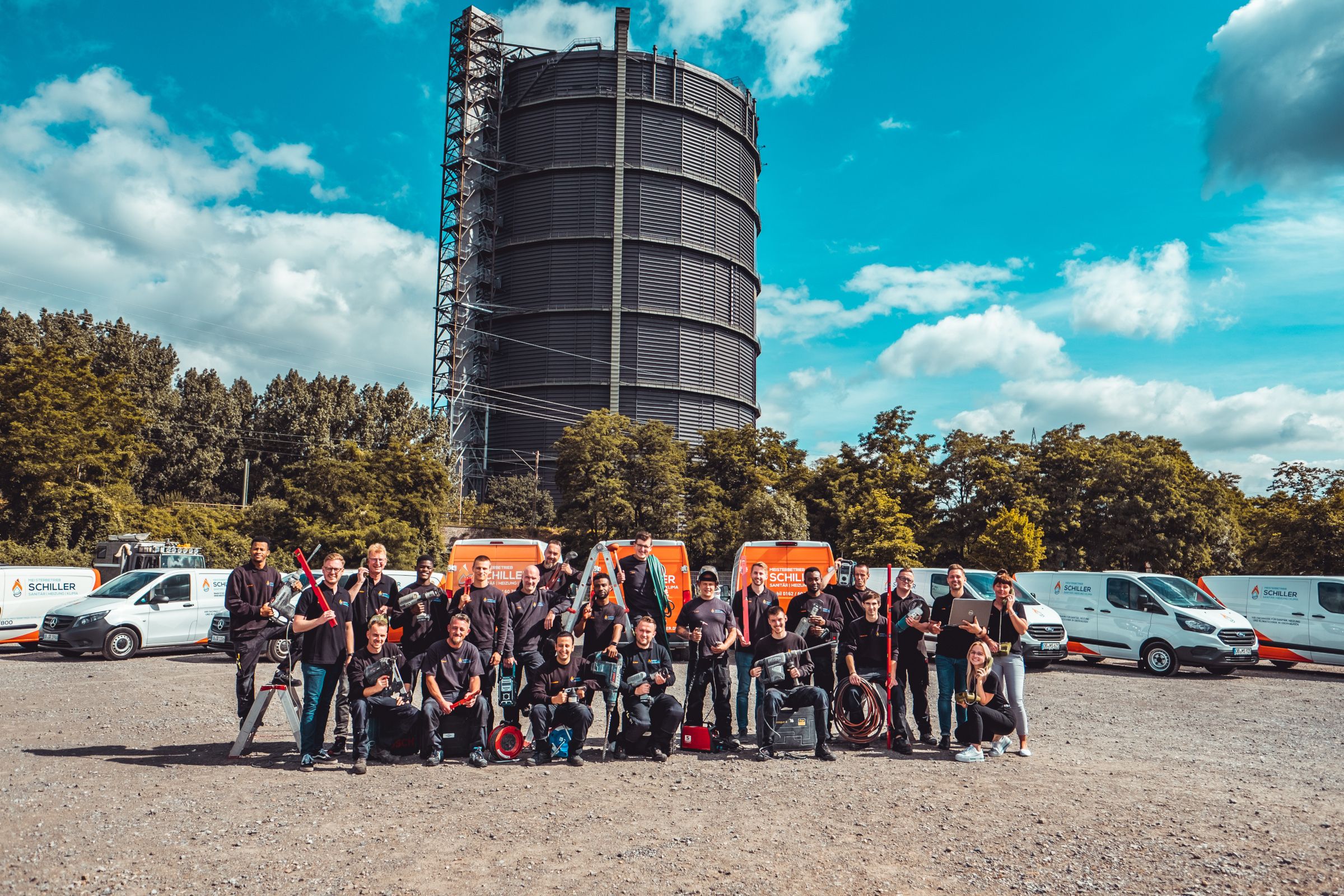 Eine große Gruppe Mitarbeitender von Schiller posiert mit Werkzeugen und Maschinen vor Firmenfahrzeugen und dem Gasometer.