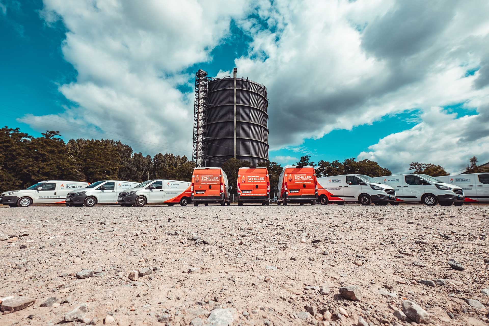 Eine Reihe von Schiller-Firmenfahrzeugen ist nebeneinander vor dem Gasometer aufgestellt, Blickrichtung auf die Rückseiten.