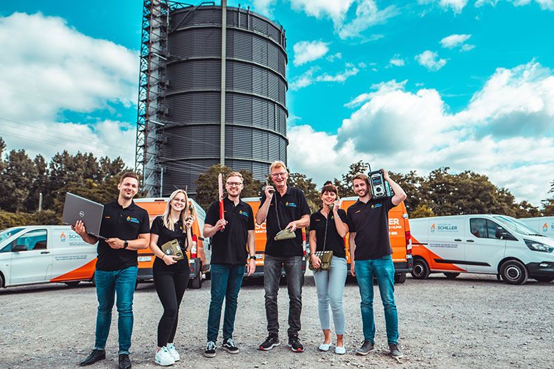 Sechs Teammitglieder posieren fröhlich mit Werkzeug vor Firmenfahrzeugen und dem Gasometer.