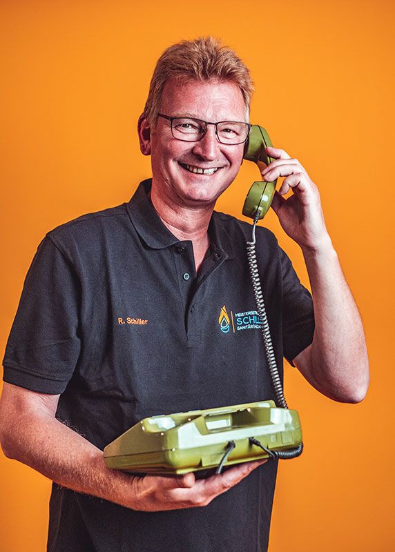 Ein lächelnder Mann in schwarzem Poloshirt mit "R. Schiller"-Stick trägt ein grünes Wählscheibentelefon vor orangefarbenem Hintergrund.